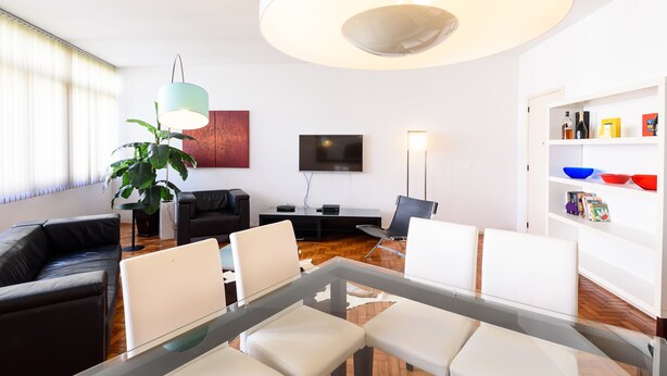 Un salon moderne avec une table à plateau de verre, des chaises blanches, un canapé noir, une grande plante en pot, un téléviseur à écran plat, une lampe sur pied et une bibliothèque avec des bols colorés.