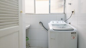 Un espace buanderie comprenant un lave-linge, un évier et une fenêtre avec des stores.