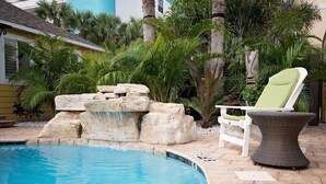 A pool area with a waterfall feature, a lounge chair, and surrounding tropical plants.