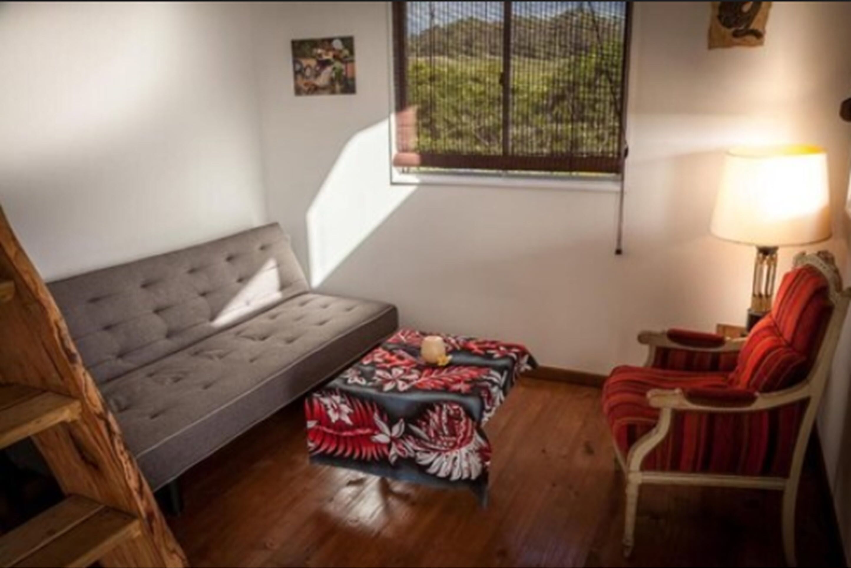 A room with a grey sofa, a red chair, a small table with a lamp, and a window with a view of greenery.