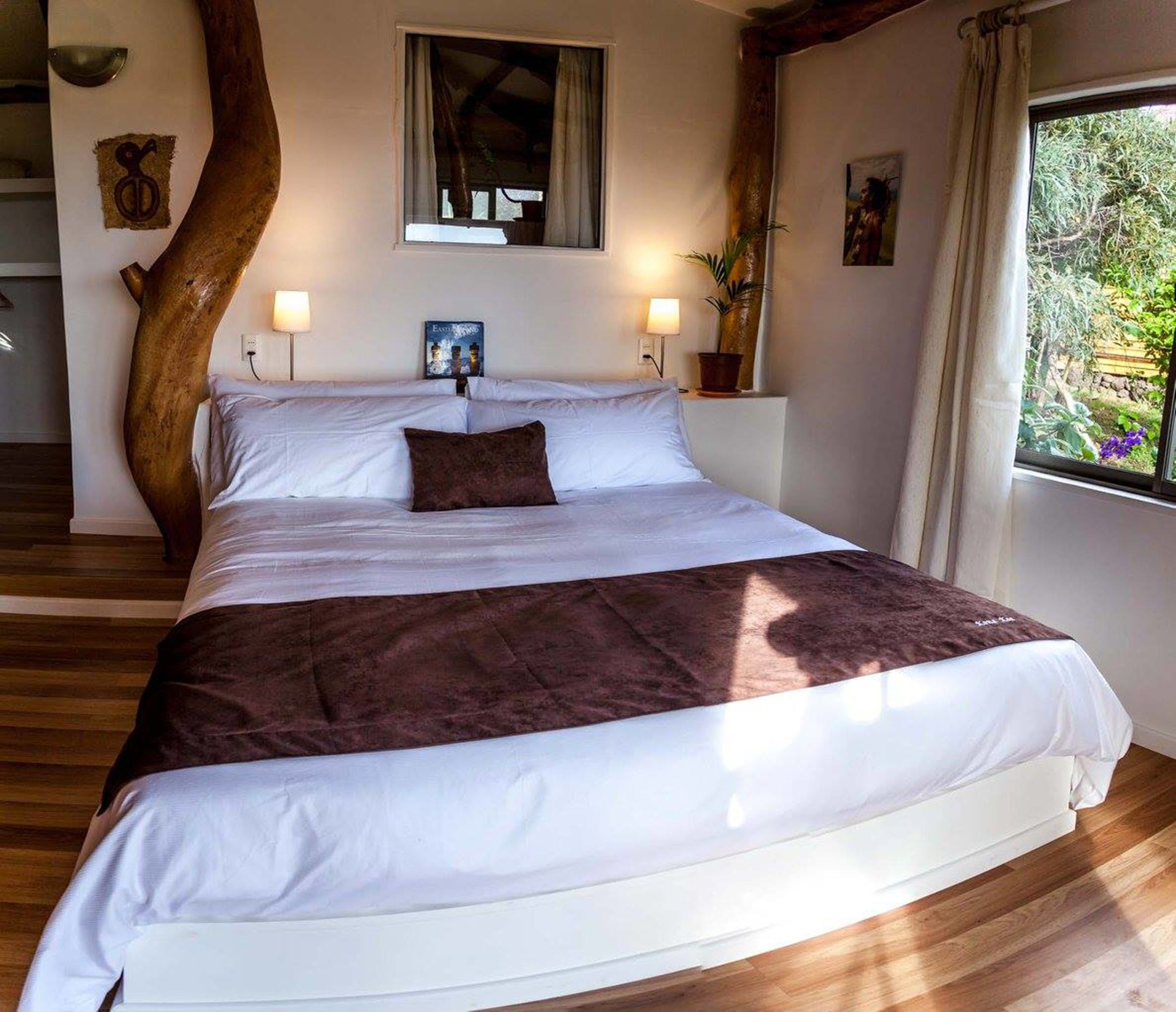 A large bed with white and brown bedding, a wooden tree trunk, a window with curtains, and a small painting on the wall.