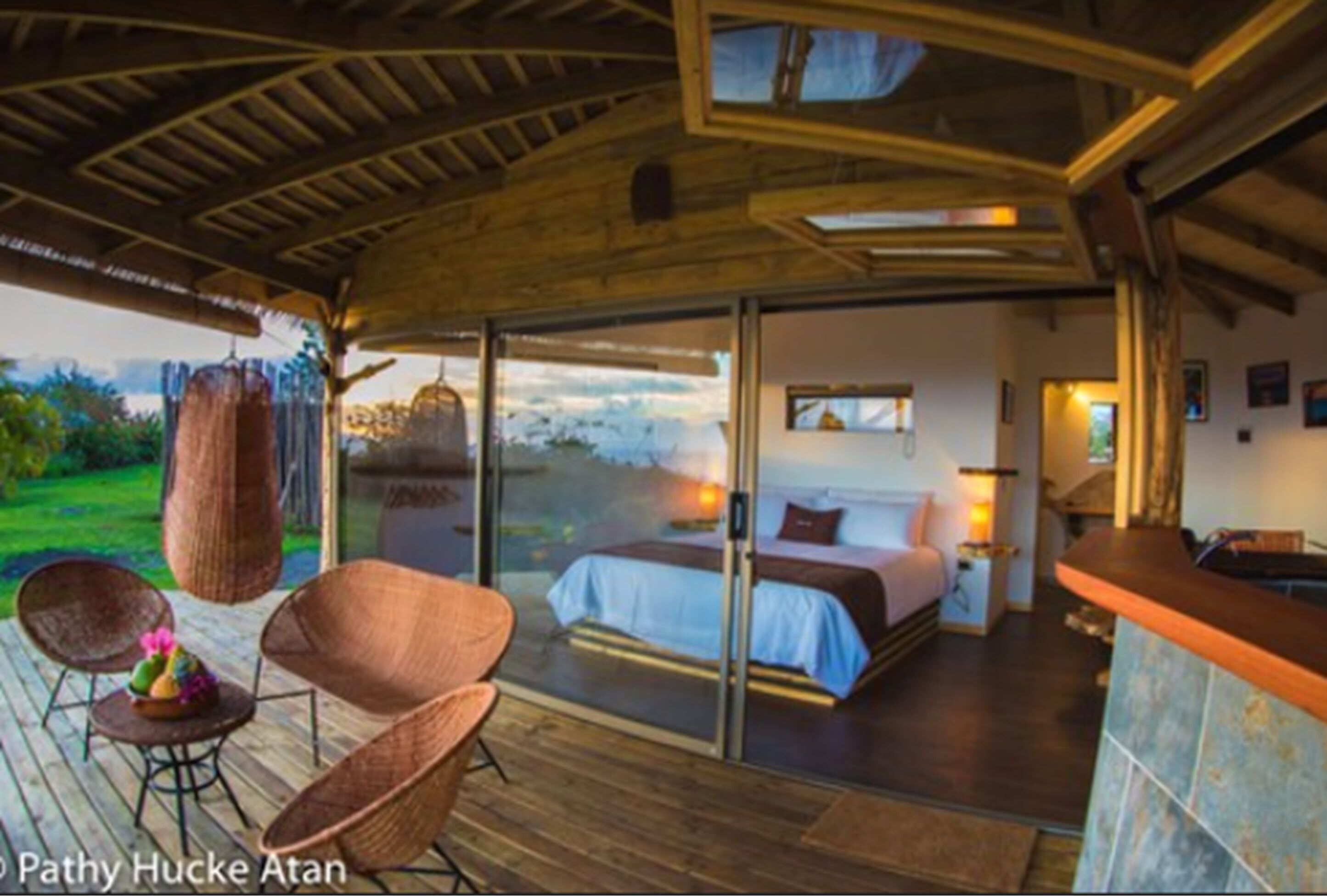 A bedroom with a large bed, a glass door leading to a balcony, wicker chairs, and a wooden ceiling.