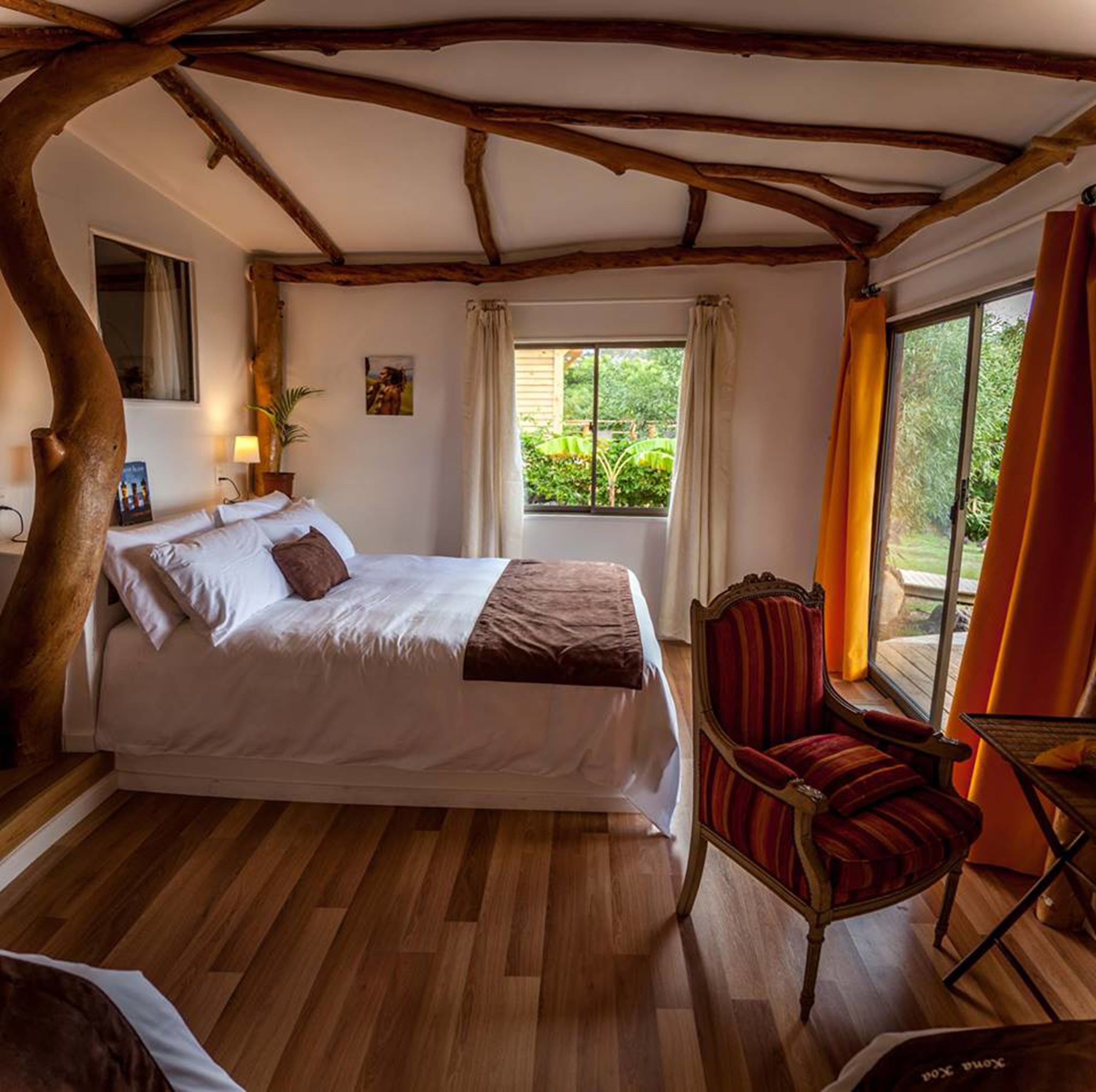 A bedroom with a wooden ceiling, a bed with white linens, a red chair, and a window with curtains.