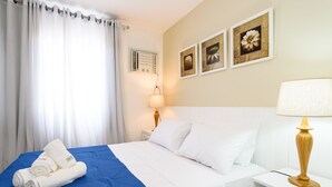 A neatly made bed with white linens and a blue accent, a bedside lamp, and wall-mounted artwork. - Omar do Rio - Apartamento CobR (Rio de Janeiro)