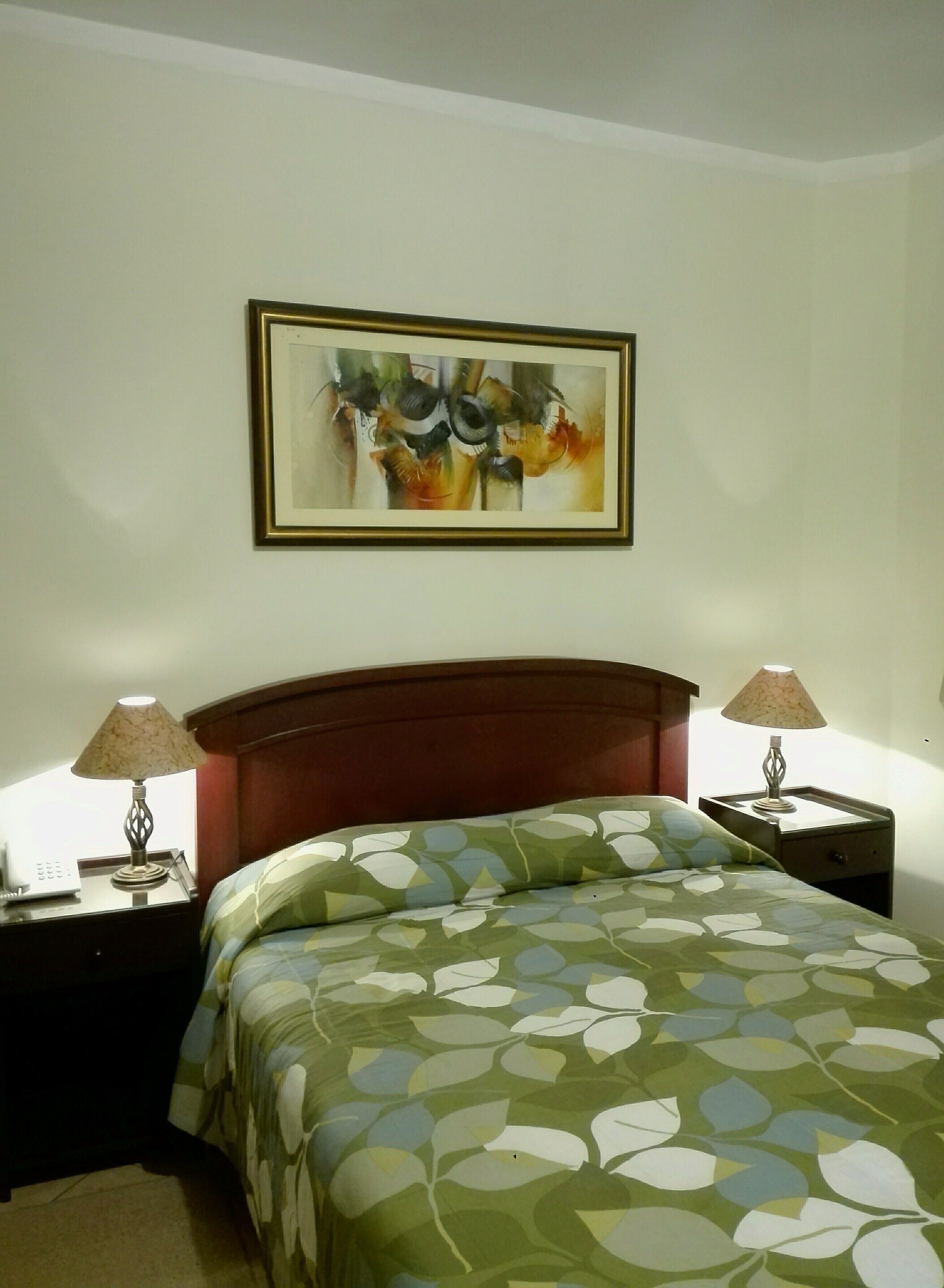 A hotel room with a bed, two nightstands, a lamp on each, and a framed abstract painting on the wall.