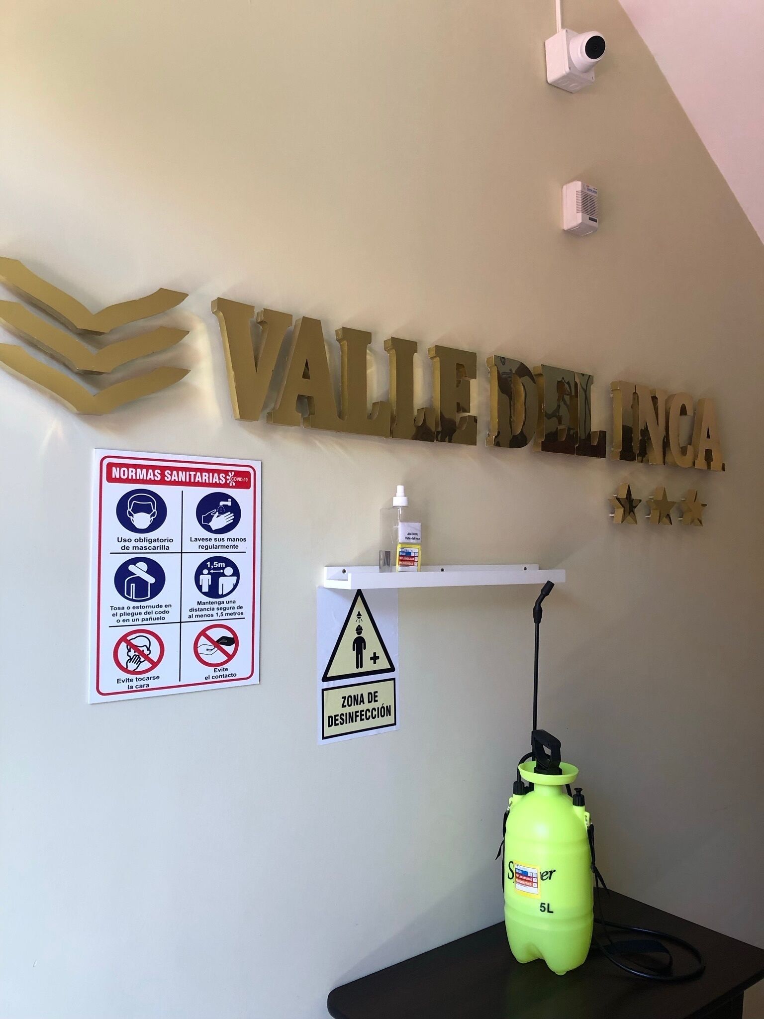 A hotel sign reading "VALLE DEL INCA" with a disinfection zone sign below it, a sanitizer dispenser, and a security camera.