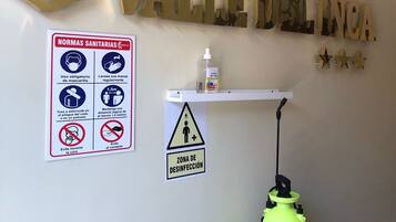 A hotel sign reading "VALLE DEL INCA" with a disinfection zone sign below it, a sanitizer dispenser, and a security camera.