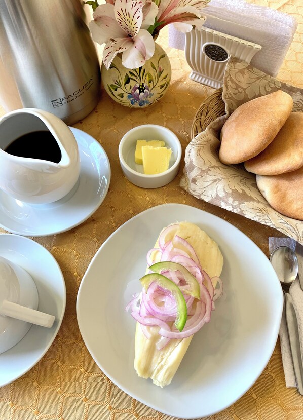 A table setting with a plate of food, a cup of coffee, a small bowl of butter, a decorative vase, and a basket of bread. - Valle del Inca (Cajamarca)