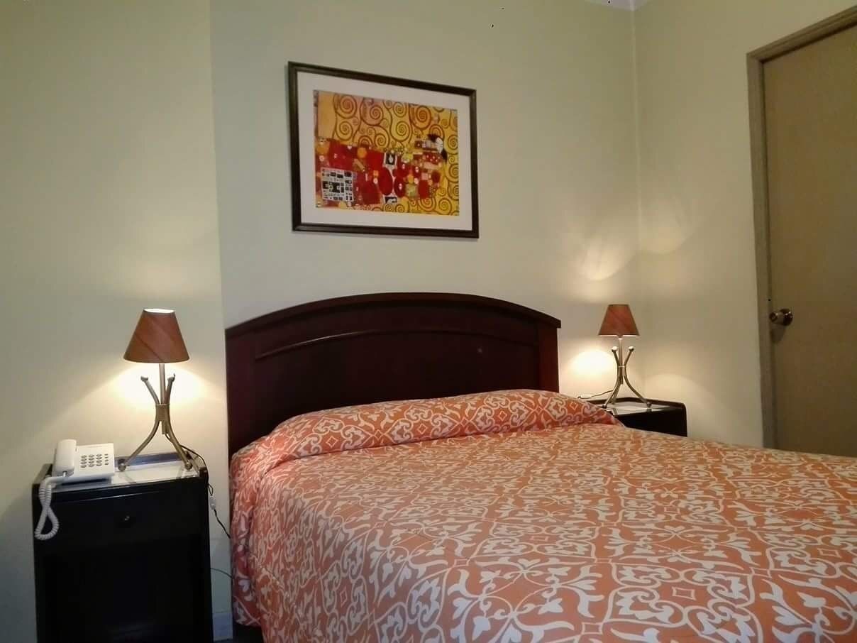 A neatly made bed with an orange patterned bedspread, two bedside tables with lamps, a framed artwork on the wall, and a closed door.