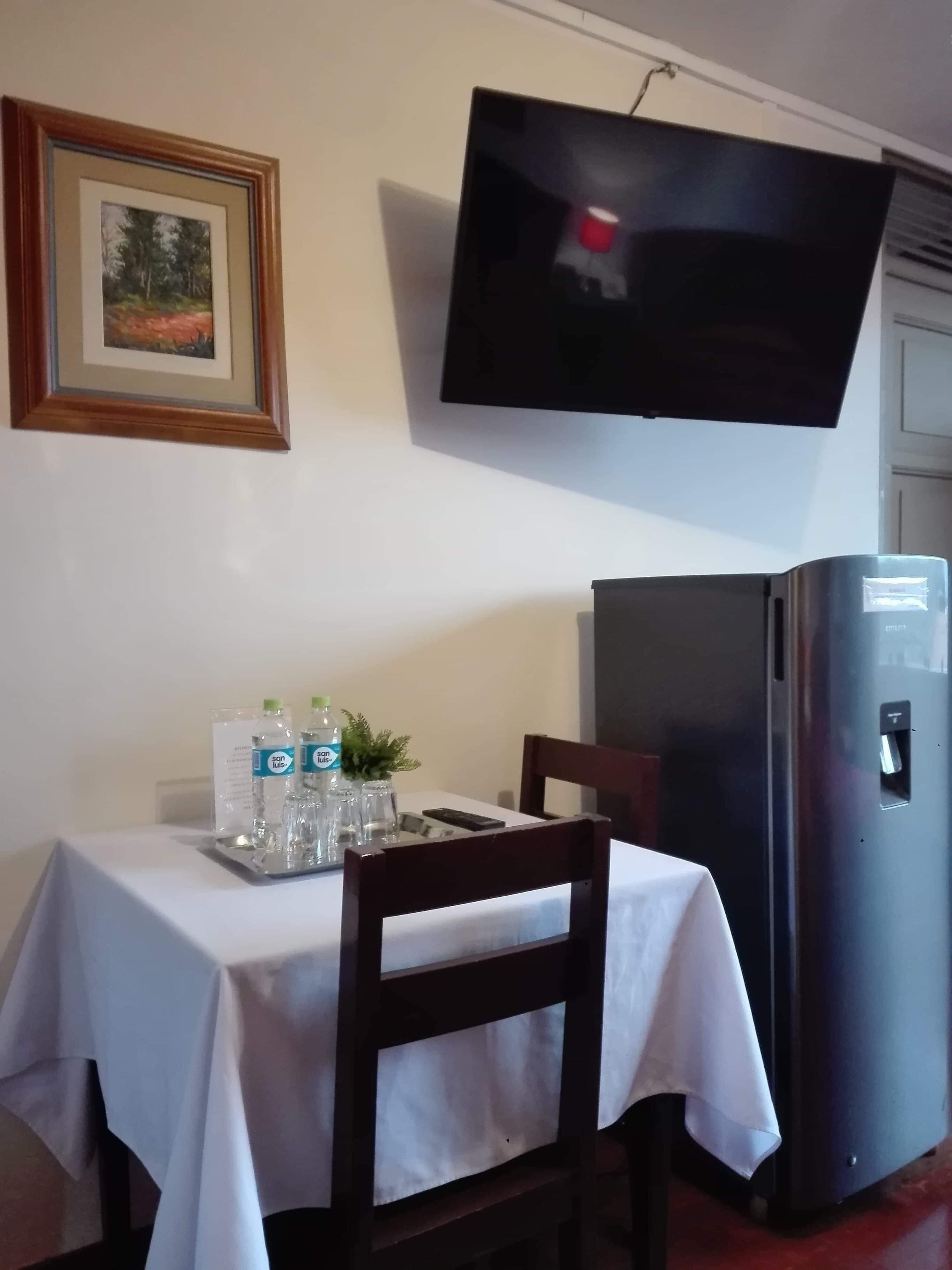 A dining area with a table set for two, a television mounted on the wall, and a refrigerator.