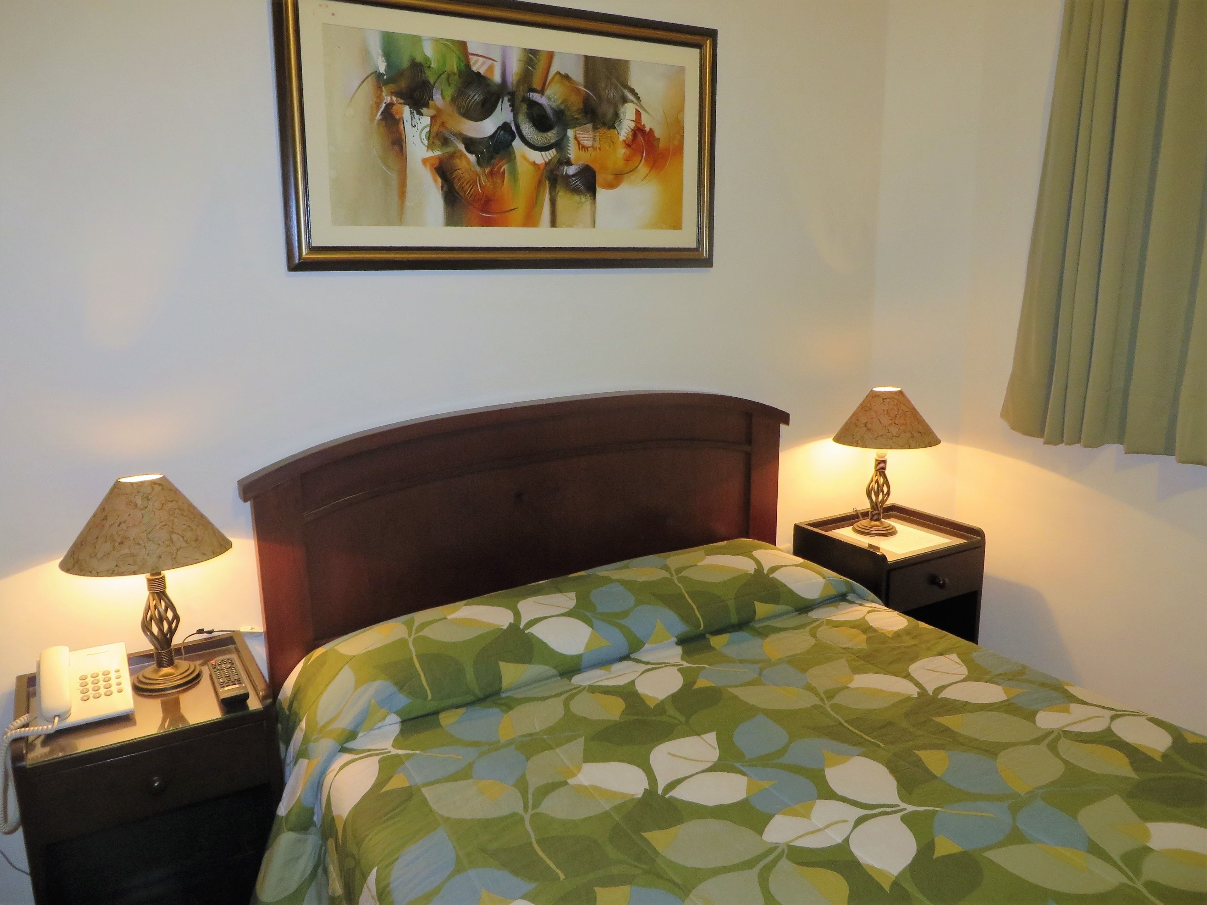 Een hotelkamer met een bed, twee bedlampen, een telefoon en een lijstig abstract schilderij aan de muur.