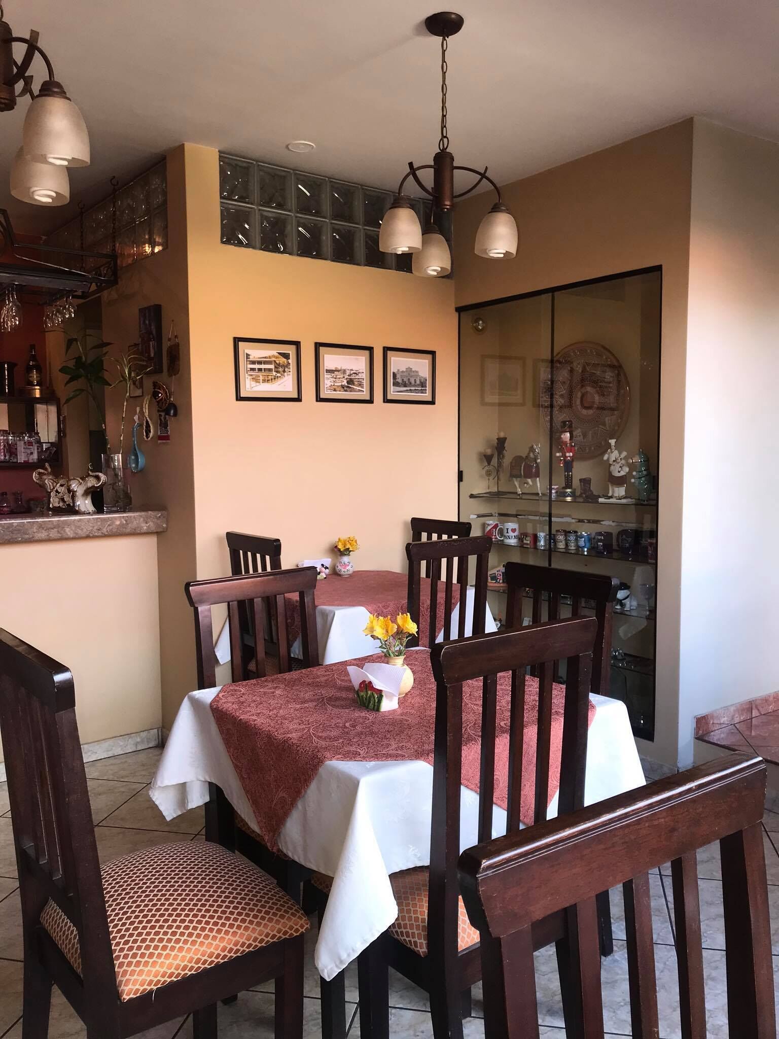 A dining area with a table set for four, surrounded by wooden chairs with cushioned seats. A chandelier hangs above the table, and there are framed pictures on the wall.