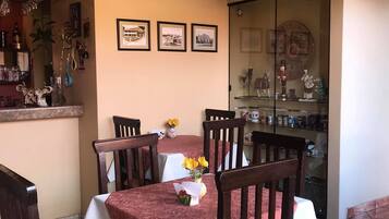 A dining area with a table set for four, surrounded by wooden chairs with cushioned seats. A chandelier hangs above the table, and there are framed pictures on the wall.