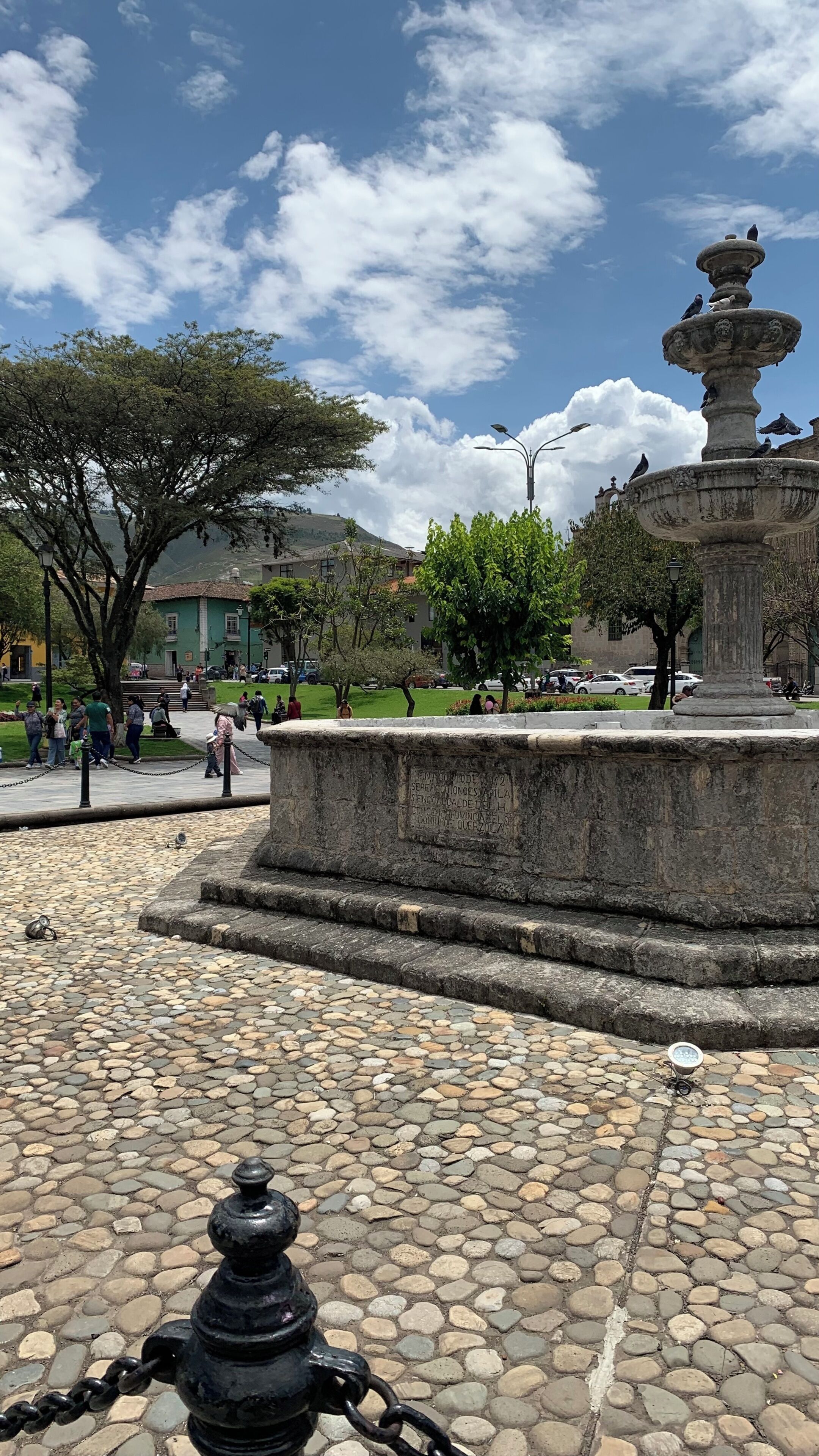 Una fuente de piedra con diseño escalonado, rodeada por una plaza adoquinada con un árbol grande y un edificio al fondo.