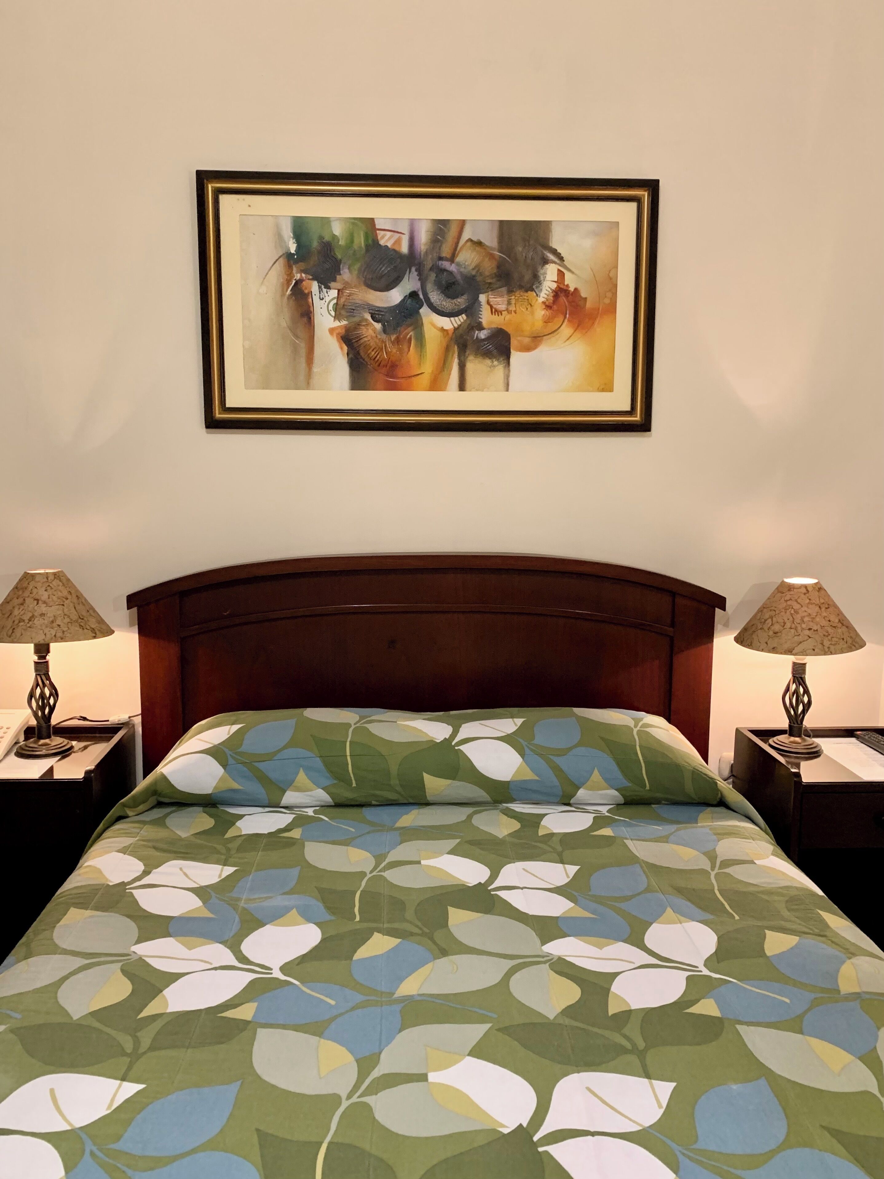 Una cama bien hecha con una colcha estampada con hojas, un cabecero de madera y un cuadro abstracto enmarcado encima. Hay dos mesitas de noche con lámparas a cada lado de la cama.
