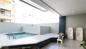 A modern indoor swimming pool with a clear view of the outside buildings, a seating area with chairs, and a small table with plants. - Omar do Rio - Apartamento FO407 (Rio de Janeiro)