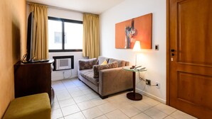 A compact living space with a sofa, a small table with a lamp, a wooden door, and a window with curtains. - Omar do Rio - Apartamento FO407 (Rio de Janeiro)