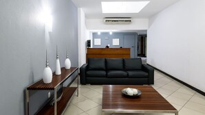 A modern hotel lobby with a black sofa, a wooden coffee table with decorative items, a console table with white lamps, and a reception desk in the background. - Omar do Rio - Apartamento FO407 (Rio de Janeiro)