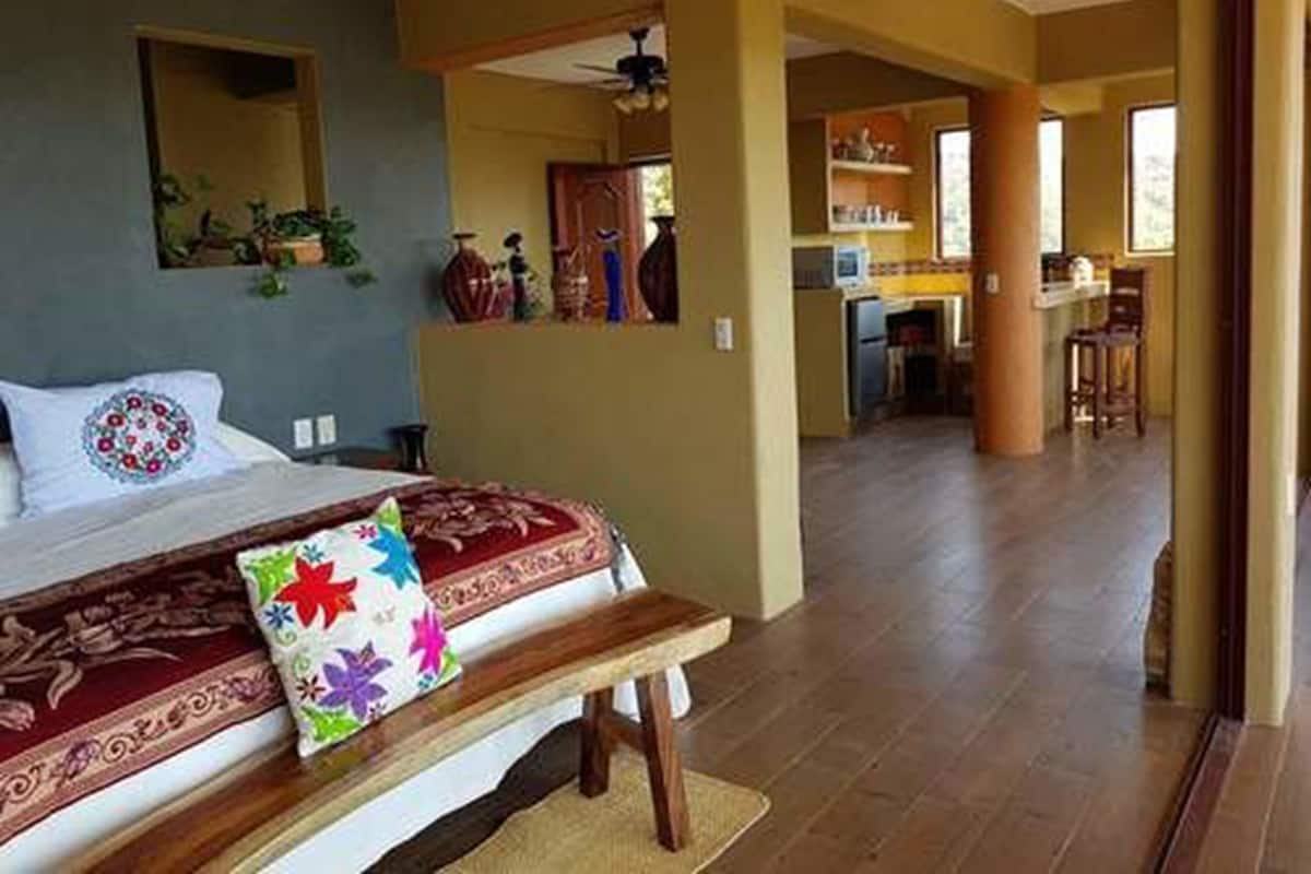 A bedroom with a bed, a bench, and a view into a kitchen area.