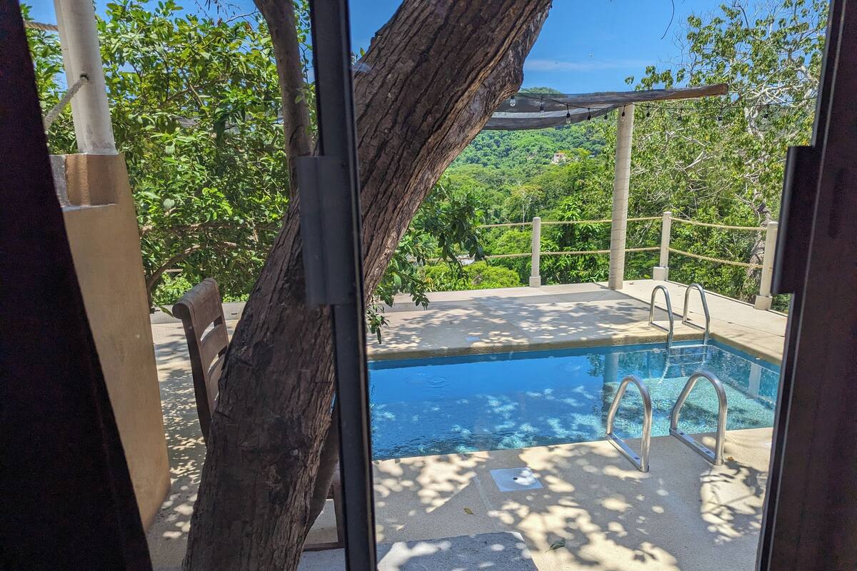 A pool area with a wooden pergola, a concrete deck, and a view of a lush green landscape.