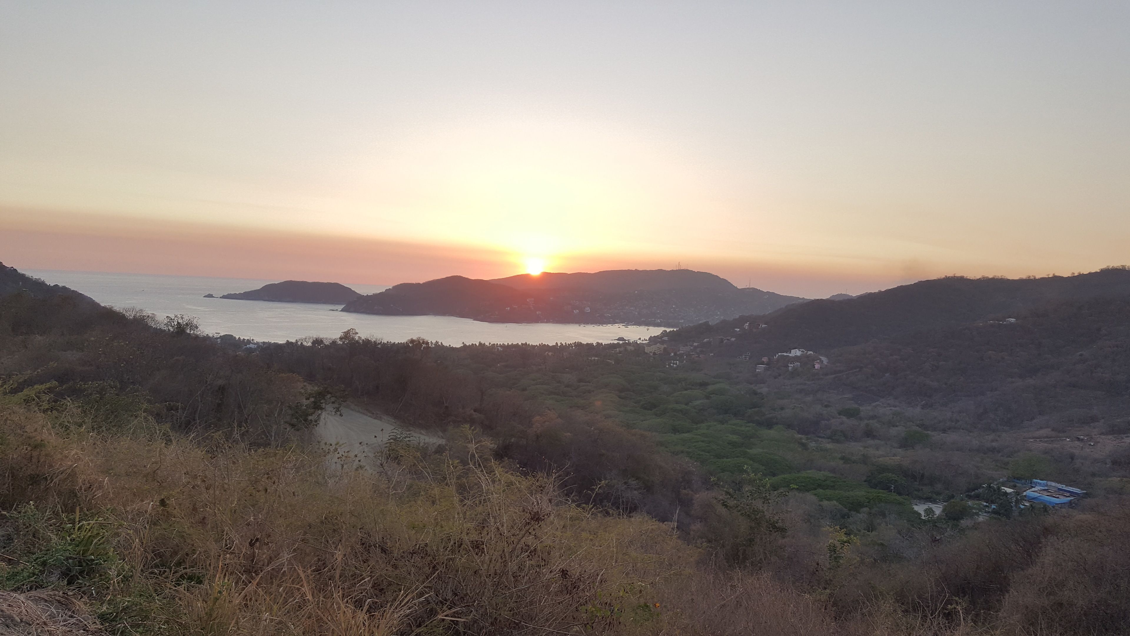 A coastal landscape with a sunset over hills and a body of water.