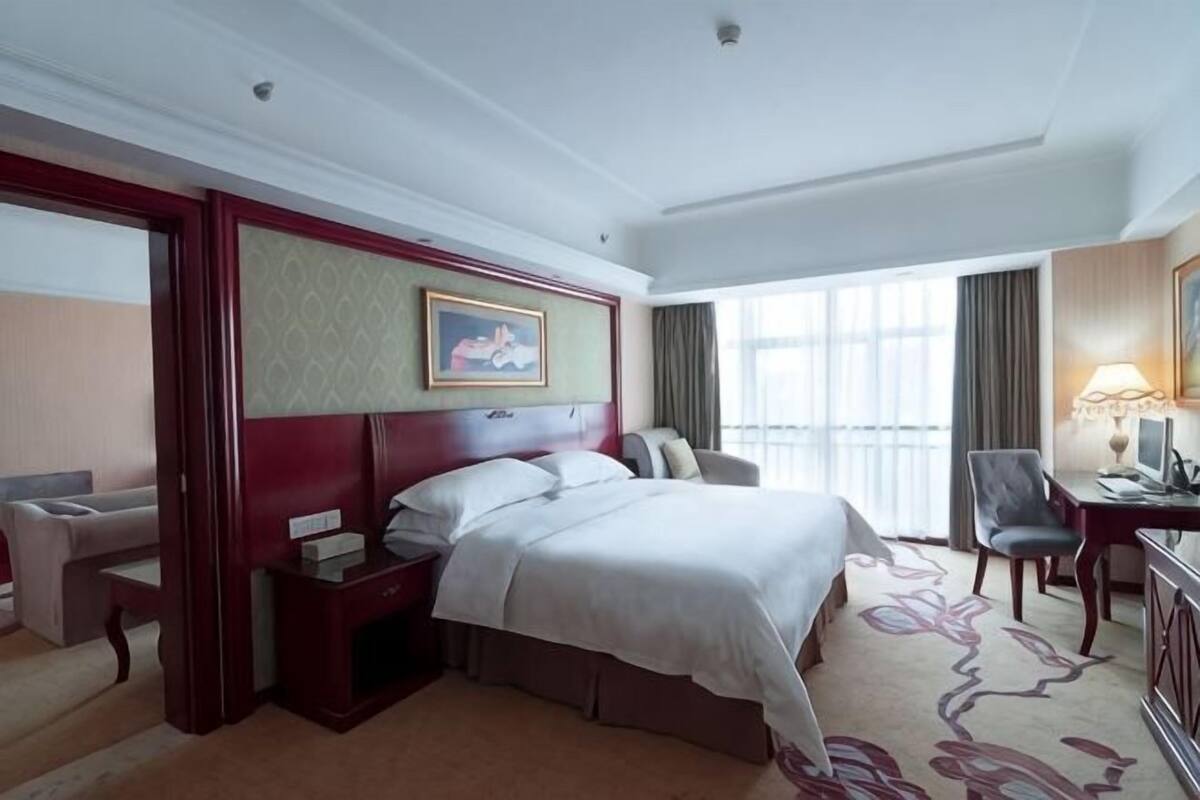 Une chambre d’hôtel avec un grand lit, un bureau, une chaise, une télévision et une fenêtre avec des rideaux.
