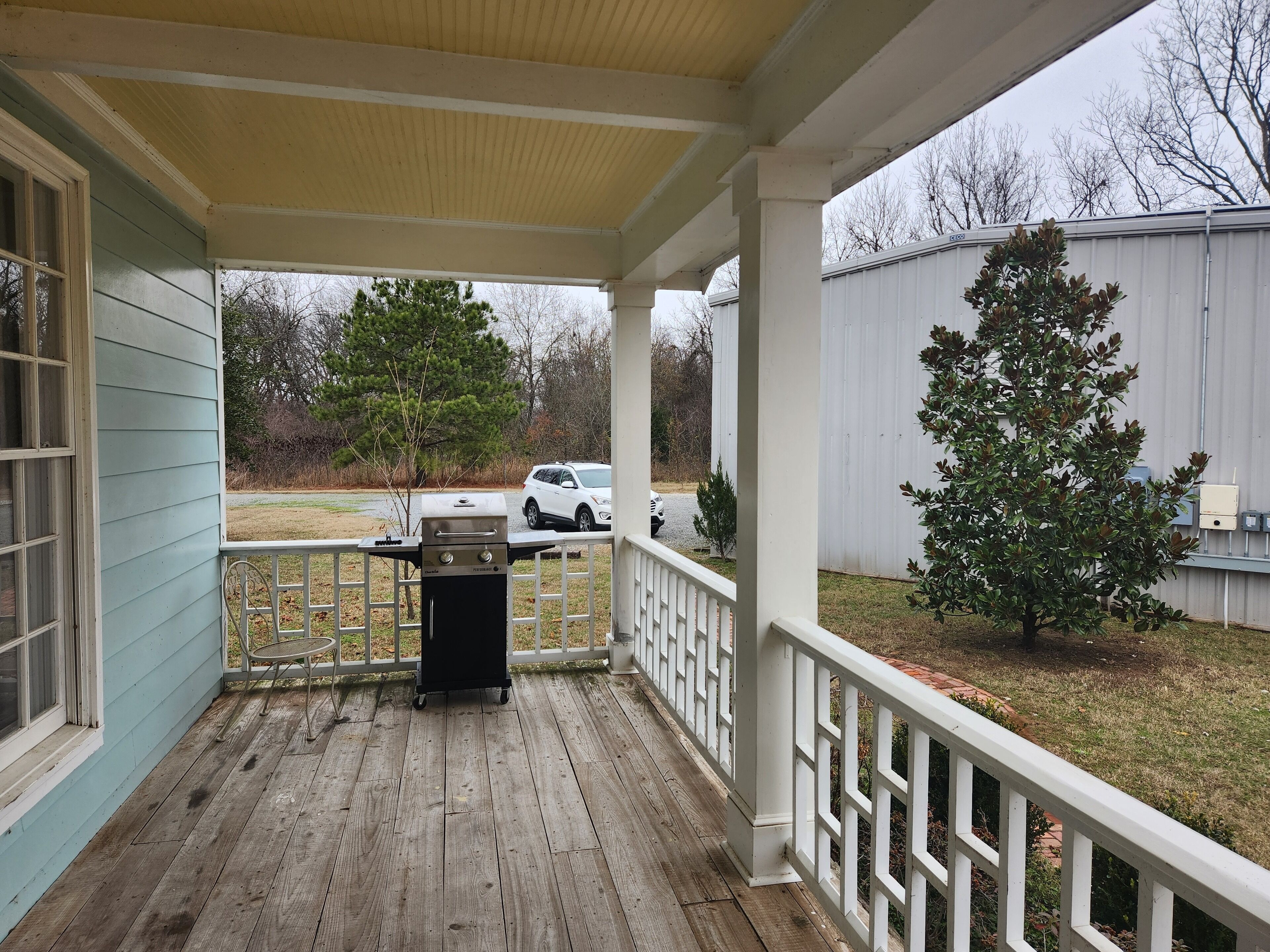 A covered porch with a grill, a white fence, and a tree.