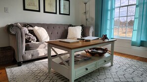 Ein Wohnzimmer mit grau gestepptem Sofa, einem Holztisch mit Schubladen, einem gemusterten Teppich und einem Fenster mit blauen Vorhängen.