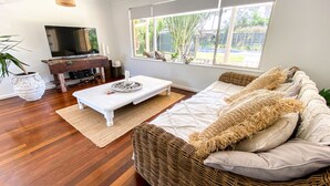 Un salon avec un canapé blanc, une table basse, un téléviseur posé sur un meuble en bois et une grande fenêtre donnant sur un paysage verdoyant.