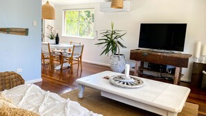 Un salon avec un canapé blanc, une table basse, un téléviseur posé sur un meuble en bois, un coin repas avec une table blanche et des chaises en osier, et une grande fenêtre donnant sur la forêt.