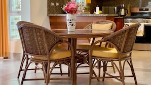 Uma sala de jantar com uma mesa redonda e quatro cadeiras de vime, um vaso com flores e uma cozinha ao fundo.