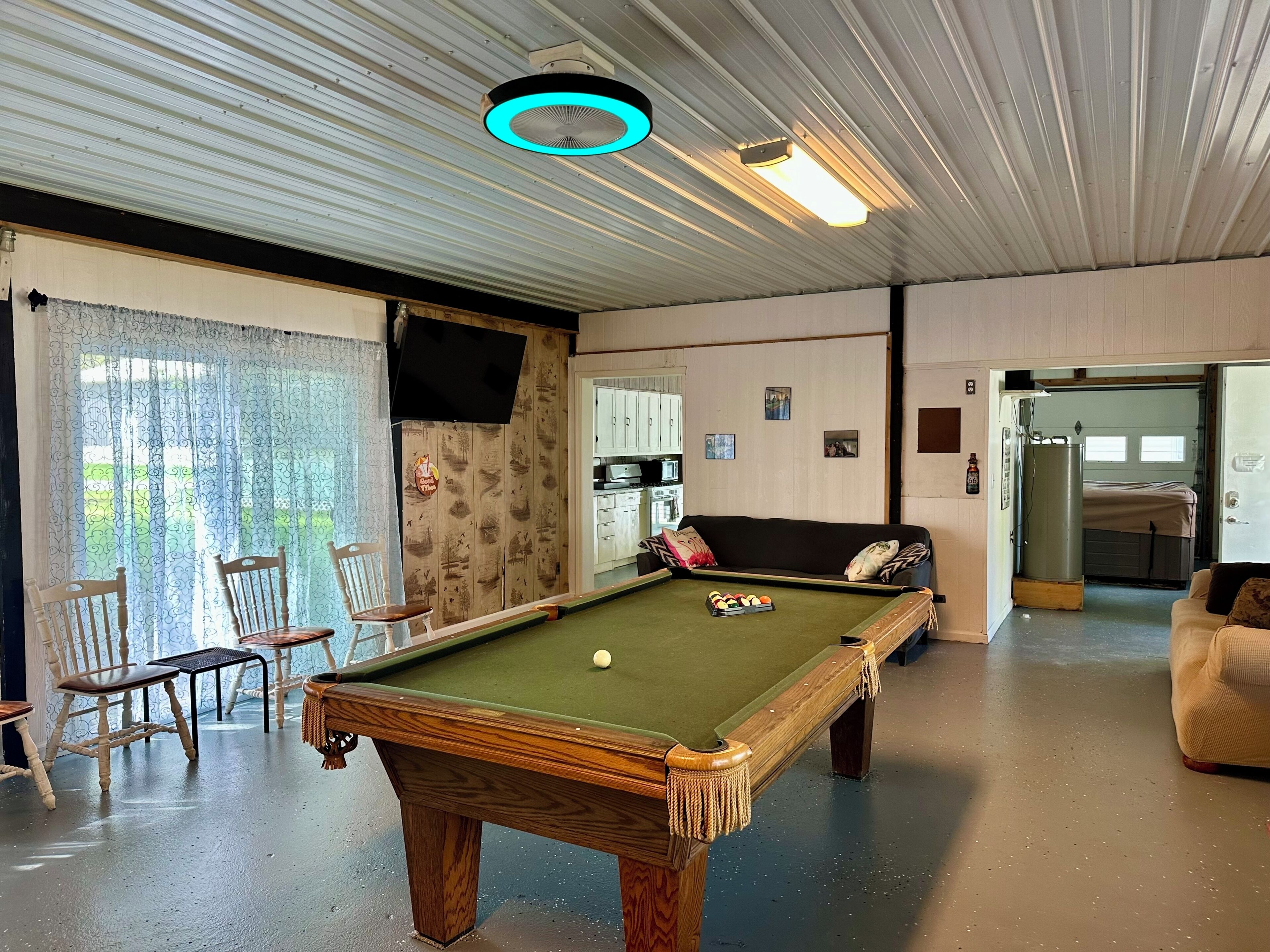 A pool table in a room with a ceiling fan, a sofa, and a kitchen area visible through an open door.