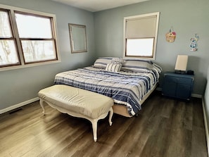 Quarto com cama, banco, cômoda e duas janelas.
