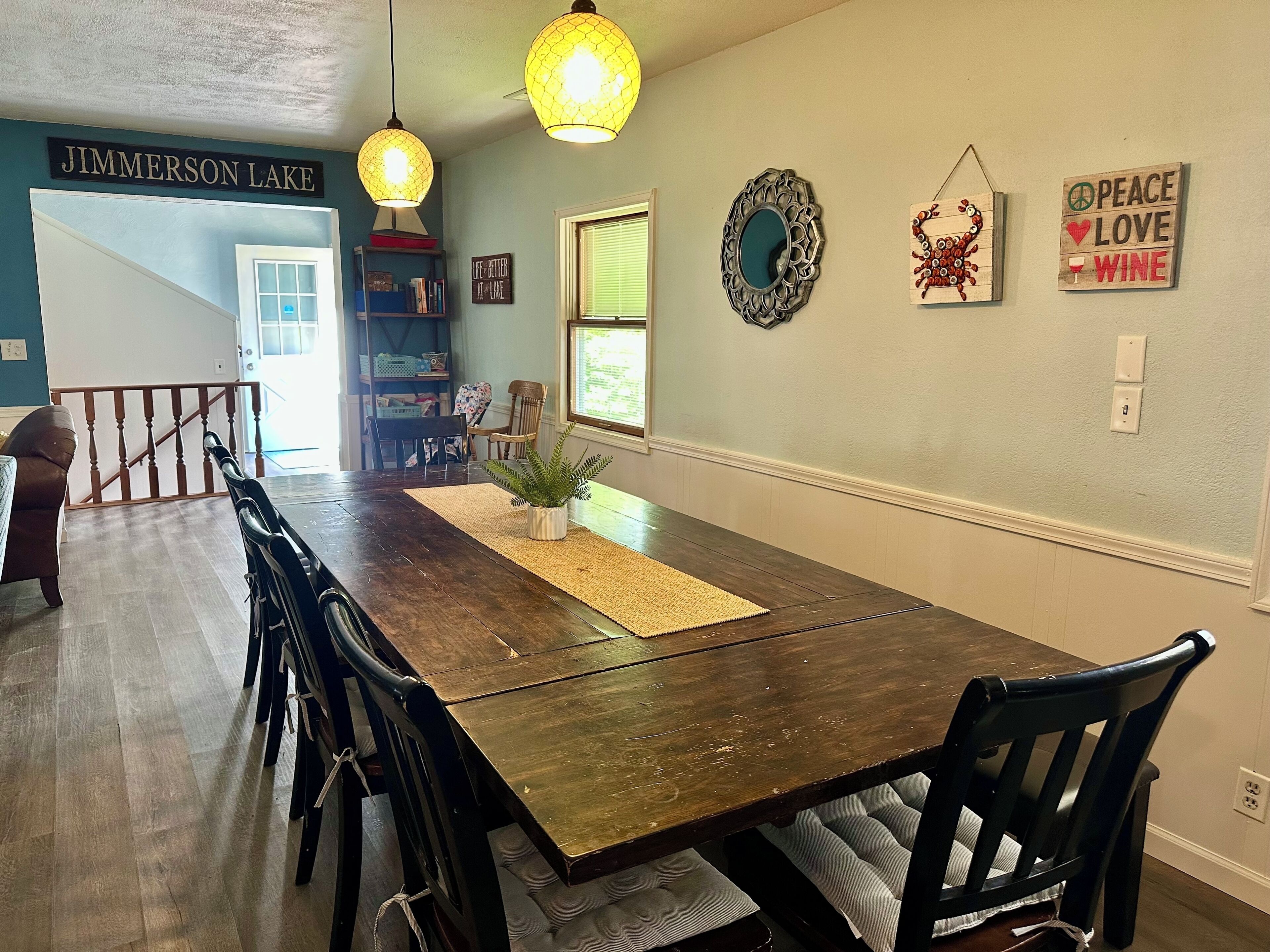 A dining area with a wooden table, chairs, pendant lights, a potted plant, and wall decorations.