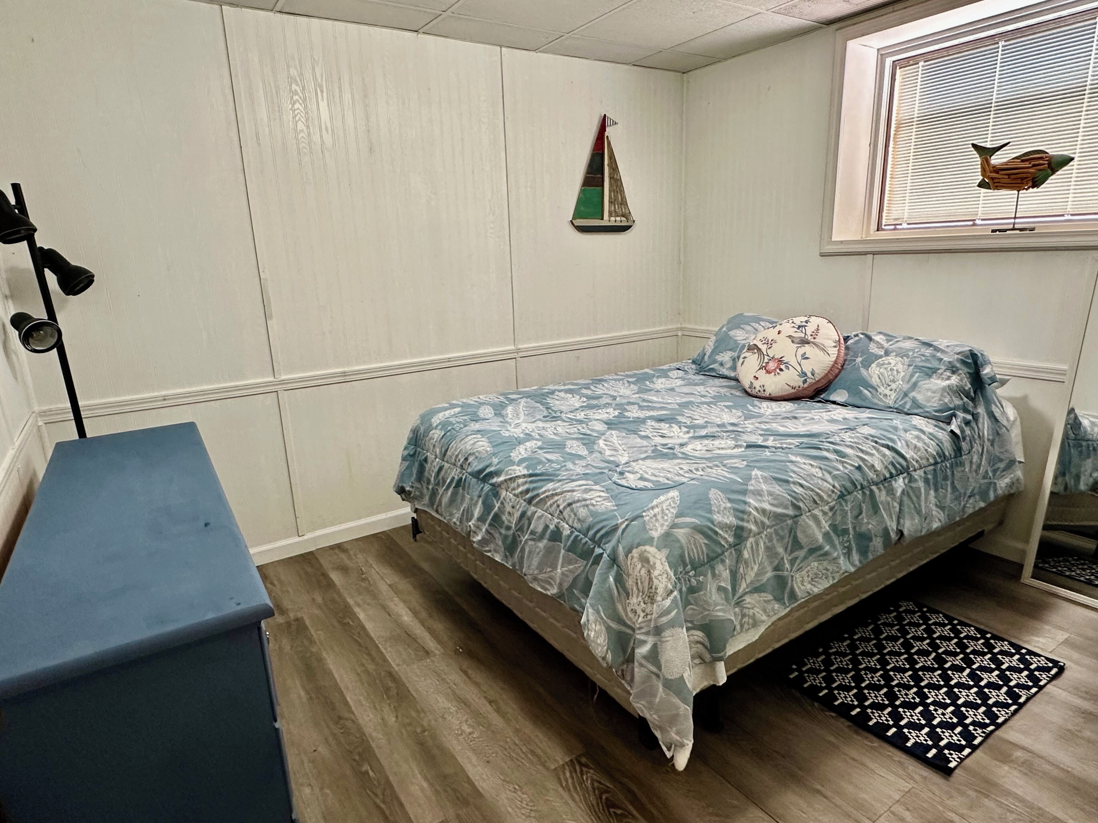 Un dormitorio con cama, escritorio, ventana con persianas y una pequeña decoración de velero.