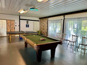 Uma mesa de bilhar com bolas, outra mesa, cadeiras e vista pela janela.
