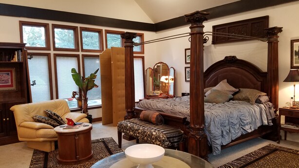 Un dormitorio amplio con cama con dosel, una zona de estar con una silla y una mesita, y ventanas grandes con persianas.