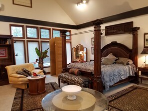 Une chambre spacieuse avec un lit à baldaquin, un coin salon comprenant un fauteuil et une petite table, ainsi que de grandes fenêtres équipées de stores.