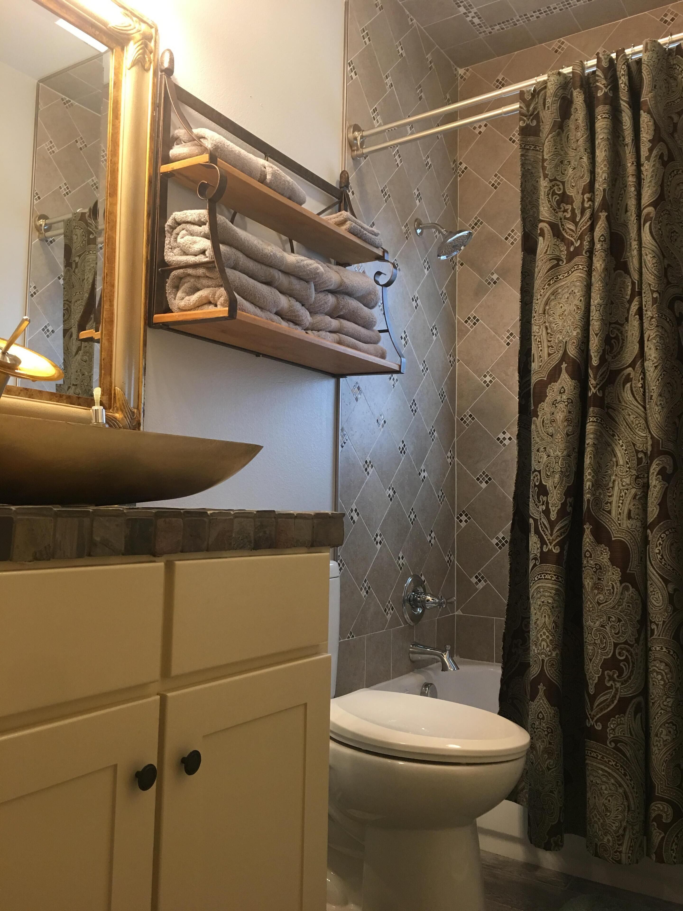 Un baño con inodoro blanco, lavamanos con espejo, toallero con toallas dobladas y cortina de ducha estampada con motivos paisley.