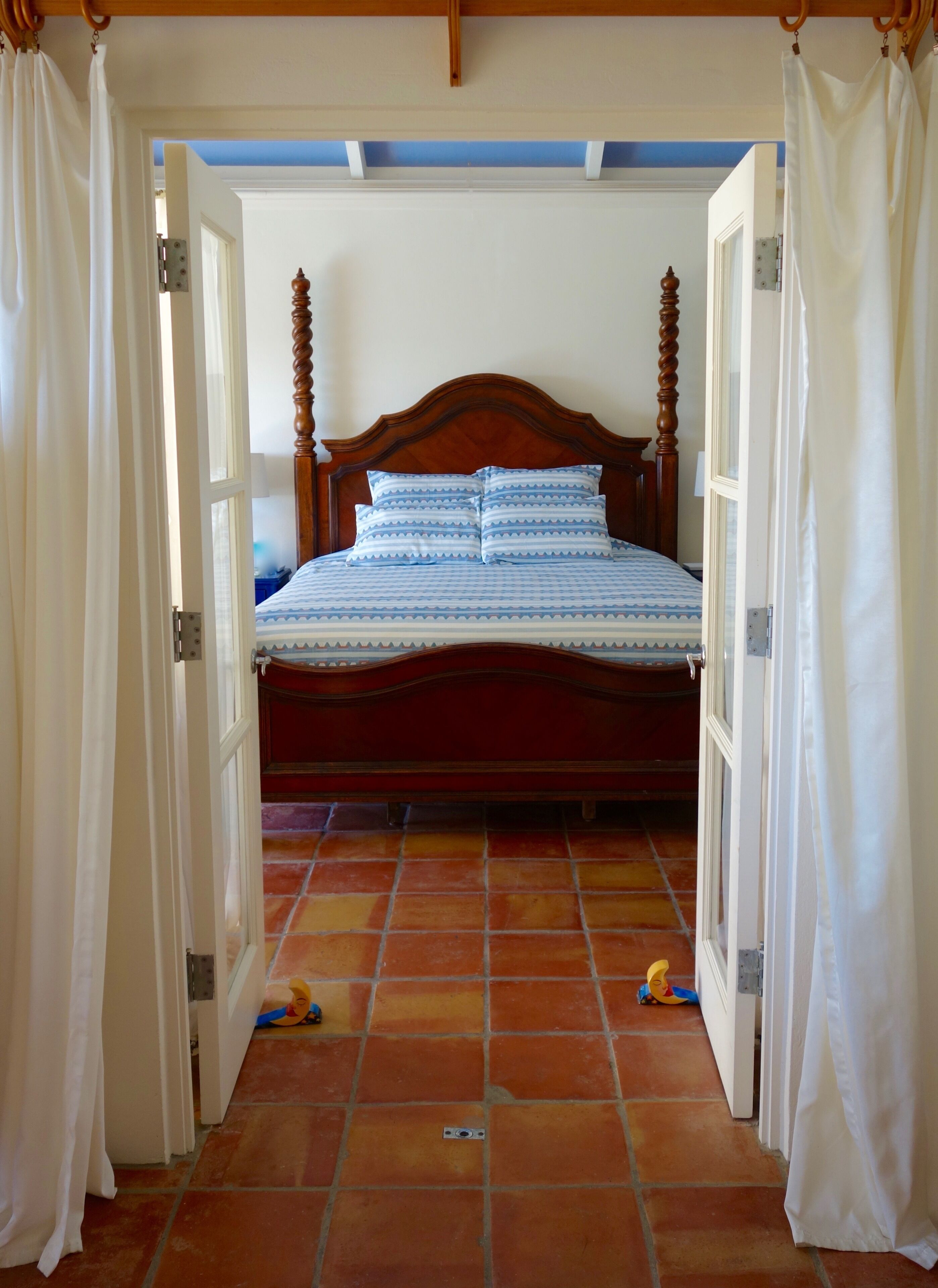 A bedroom with a wooden bed frame, white curtains, and a tiled floor.