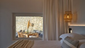 Un dormitorio con un ventanal grande, una pared de piedra, una zona de comedor con sillas y una mesa, y una cama con cabecero.