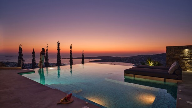 Piscina infinita con vistas al atardecer, pared de piedra y zona de descanso con cojín.