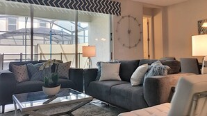 Una sala moderna con un sofá gris, una mesa de centro de cristal y una planta en maceta. Hay un reloj grande en la pared y una lámpara al lado del sofá.