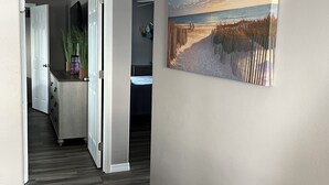 A hallway with a wooden floor, a painting of a beach scene, and a door leading to a room with a dark-colored dresser and a plant. - Beautiful Home in Ft Myers Beach. (Fort Myers Beach)