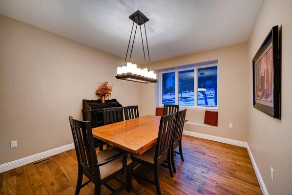 A dining room with a wooden table and chairs, a chandelier, a window with a view of a snowy landscape, and a framed picture on the wall.