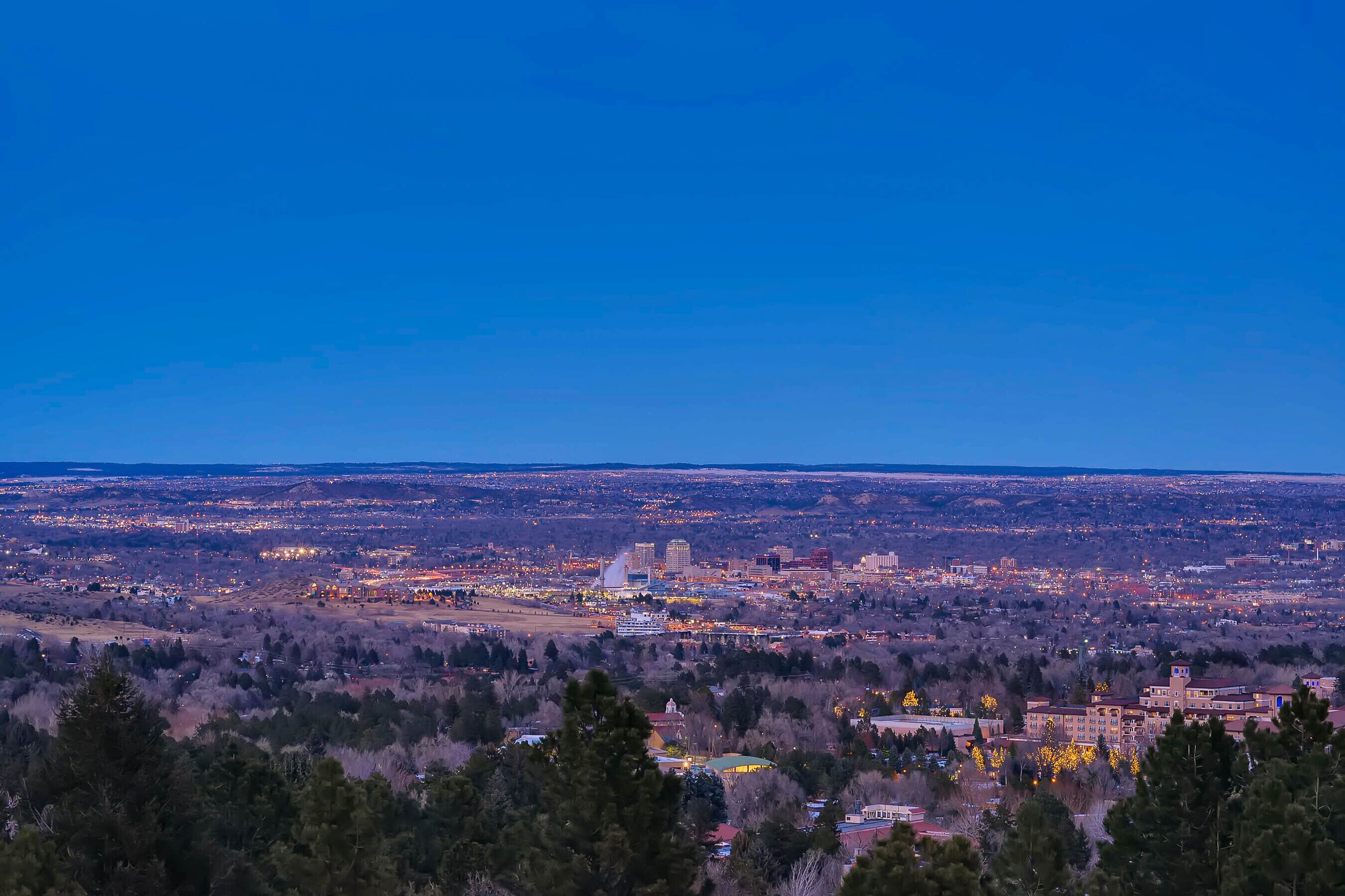 A panoramic view of a cityscape at dusk with illuminated buildings and a distant mountain range.