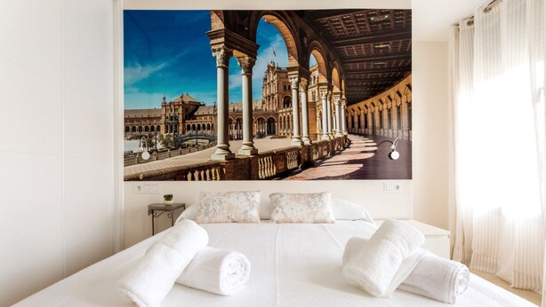 Une chambre d’hôtel avec un grand lit, du linge de lit blanc, deux oreillers et un tableau représentant un bâtiment historique.
