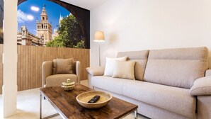 Un salon avec un canapé beige, une table basse en bois sur laquelle se trouvent un bol et une télécommande, une lampe, et une fresque murale représentant un paysage urbain.