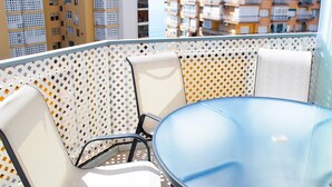 Un balcón con una mesa de vidrio redonda y sillas blancas, con vista a un paisaje urbano de edificios altos.