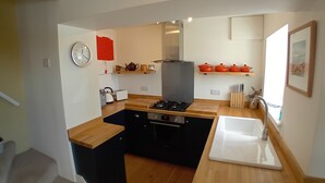 Cocina pequeña con estufa a gas, un lavamanos y encimeras de madera. Hay un reloj en la pared y un estante rojo con utensilios de cocina.